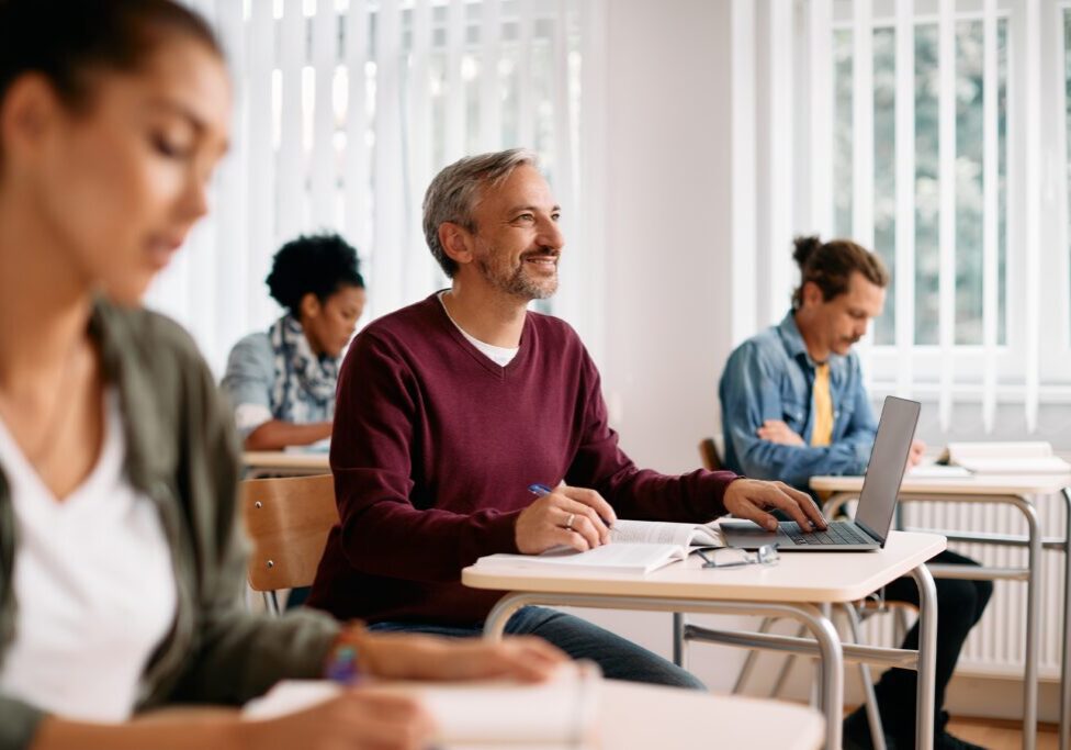 Happy,Mature,Student,Writing,Notes,And,Using,Laptop,While,Learning